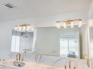 Bright modern bathroom featuring a large mirror, gold fixtures, and marble surfaces.