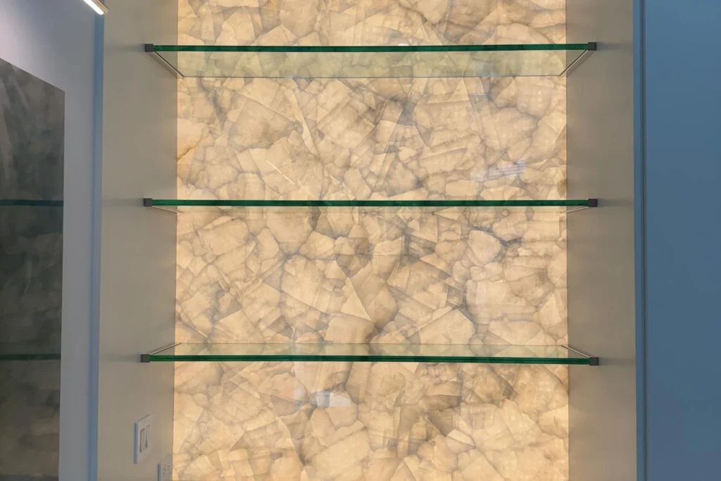 Glass shelves with polished chrome U-channels over a backlit stone wall by ACG Glass & Metals in West Palm Beach